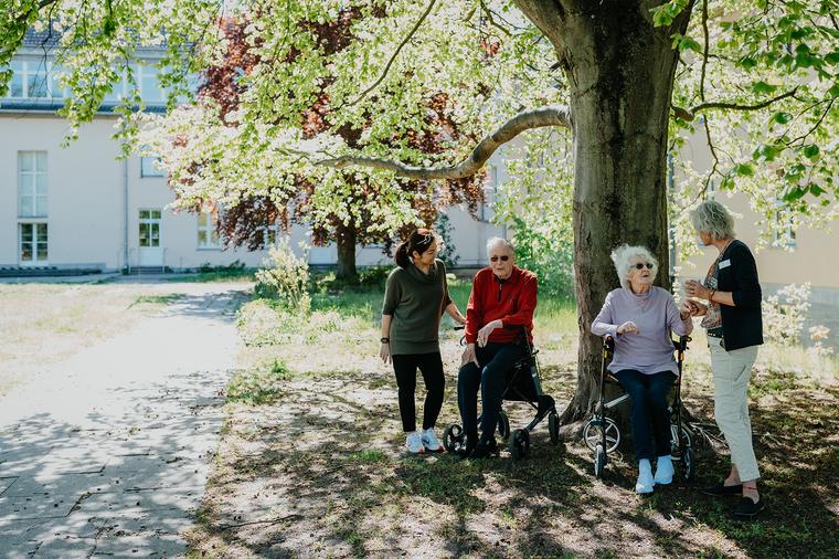 zwei Mitarbeiterinnen und zwei Bewohnerinnen sitzen im Freien unter einem schönen Baum im Schatten